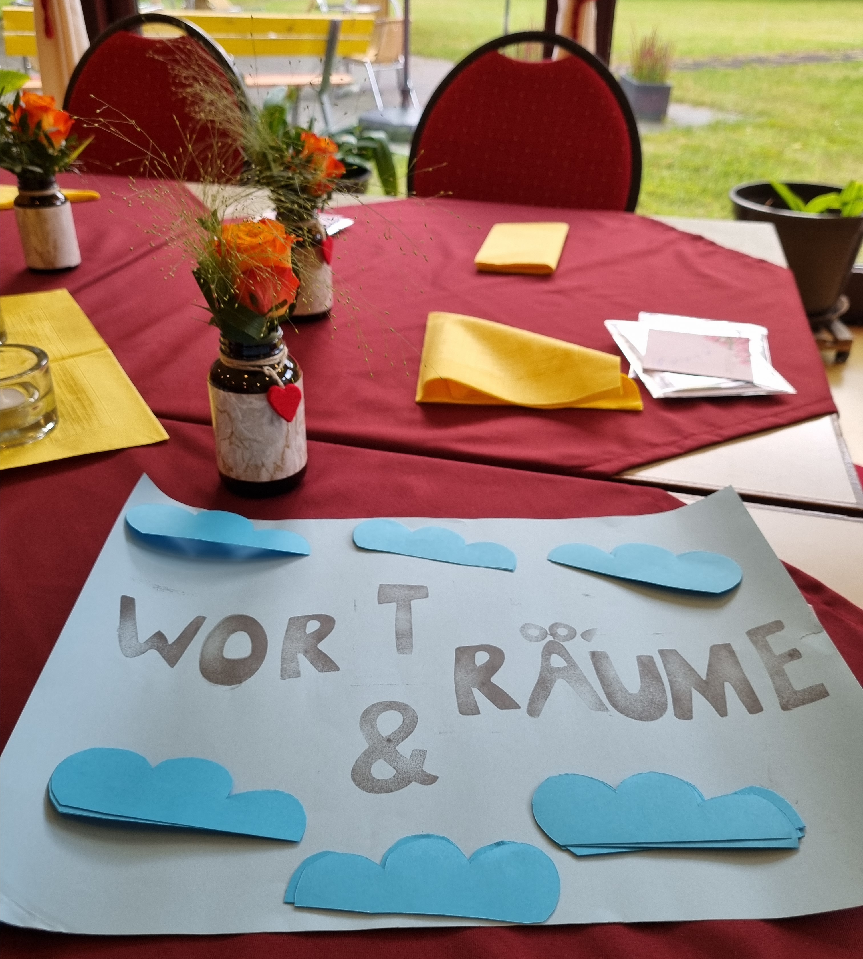 Tischdeko mit Blumen und Schild "Wort & Räume" auf farbigen Servietten in einem hellen Raum mit Blick in den Garten.