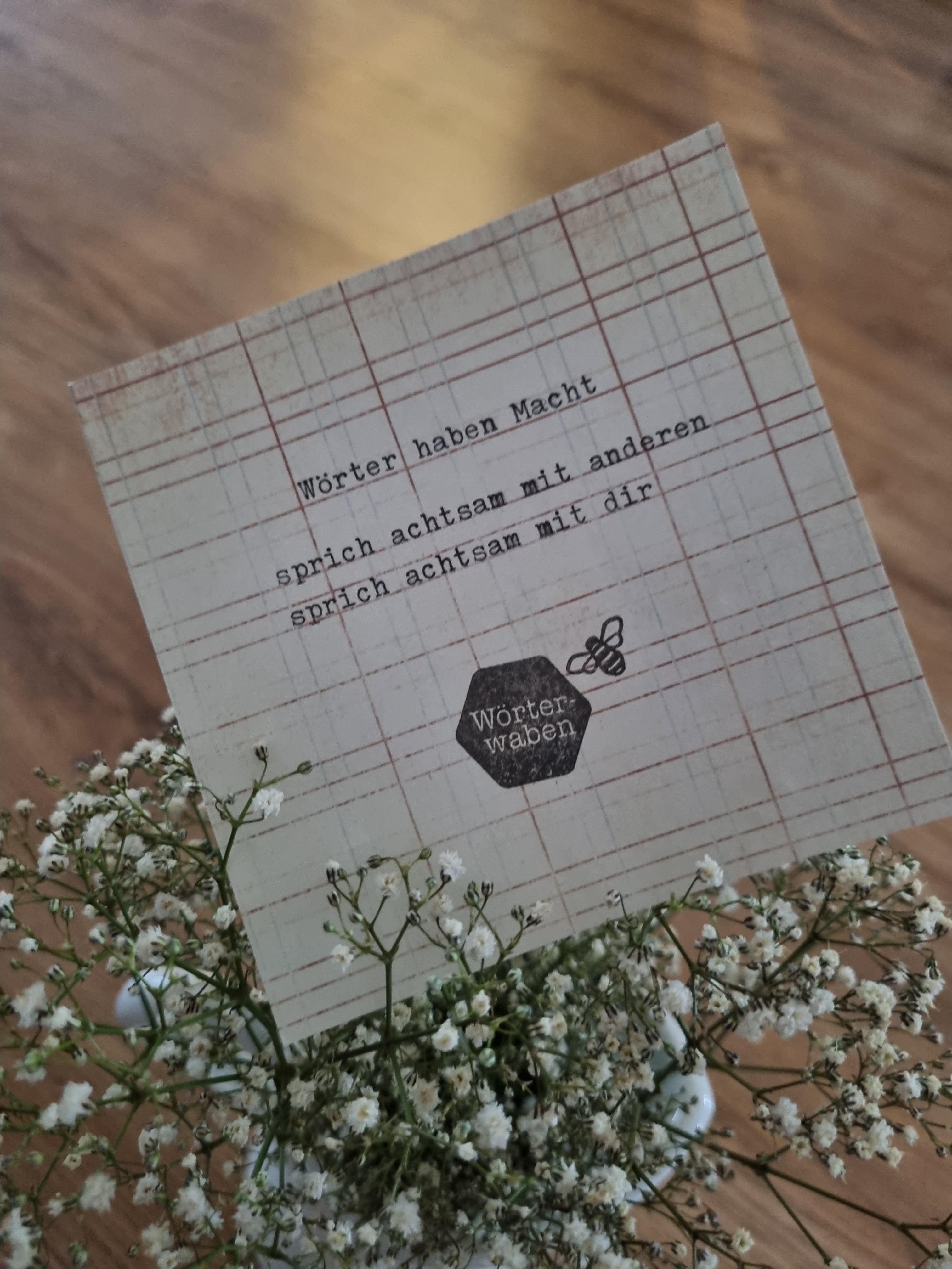 Karte mit dem Logo „Wörterwaben“ steckt in einem Strauß weißer Blüten auf einem Holzboden.
